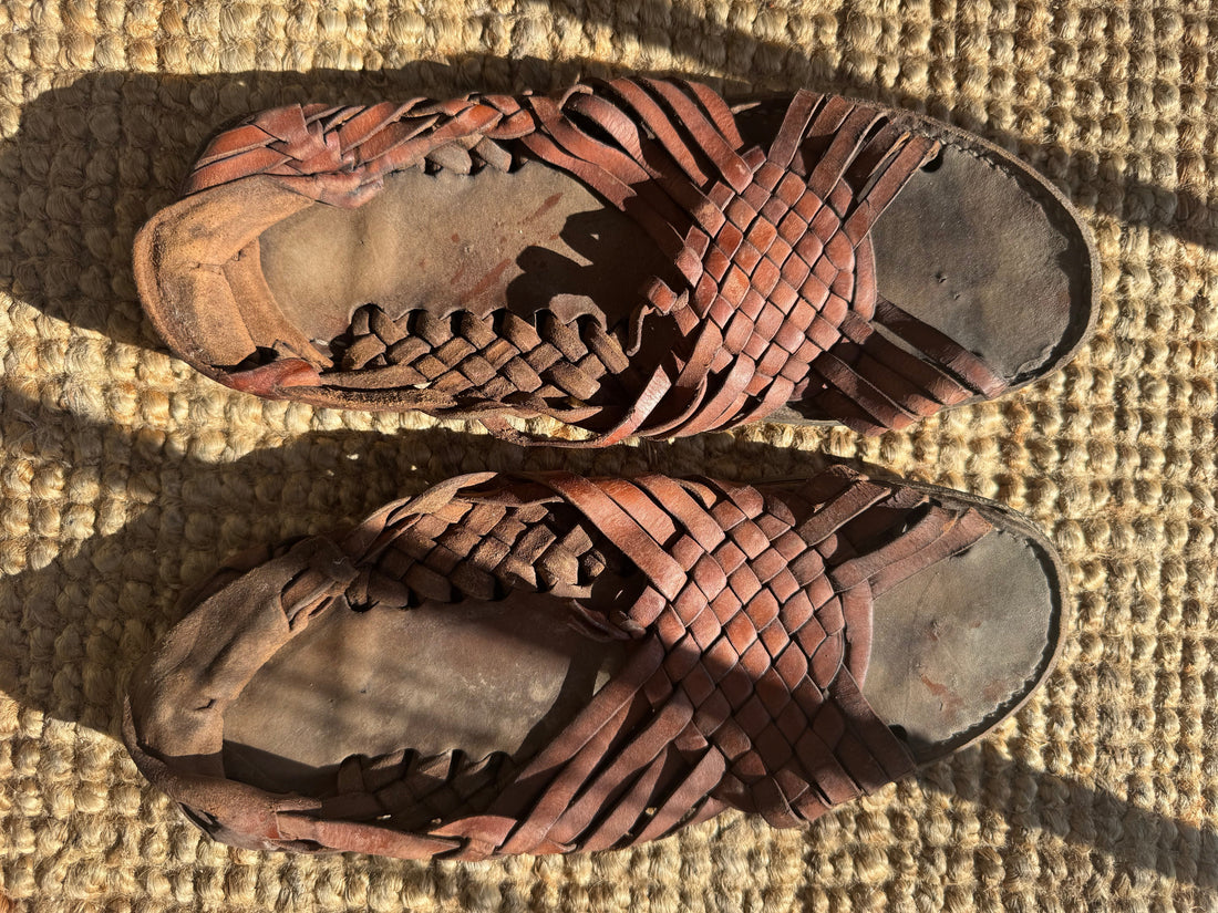 A pair of worn, brown leather Huaraches with woven straps placed on a textured, tan-colored rug.