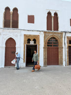 A man in a green shirt and jeans carries a brown leather suitcase while walking in front of a white building with brown wooden doors and arches. Another person stands near the left doorway carrying bags.