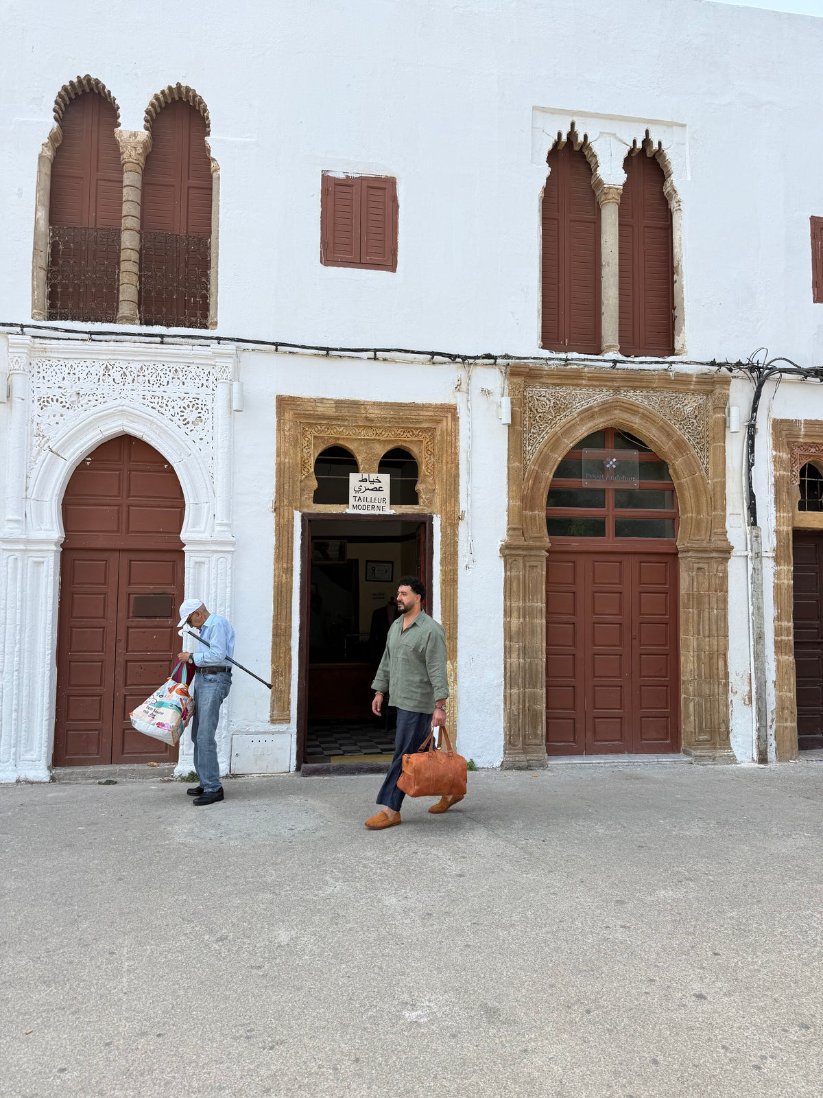 A man in a green shirt and jeans carries a brown leather suitcase while walking in front of a white building with brown wooden doors and arches. Another person stands near the left doorway carrying bags.