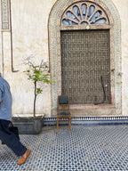 A person in a blue shirt and dark trousers walks past a traditional Moroccan-style window with a metal grille, a small wooden chair, and a potted tree on a blue-and-white patterned tile floor.