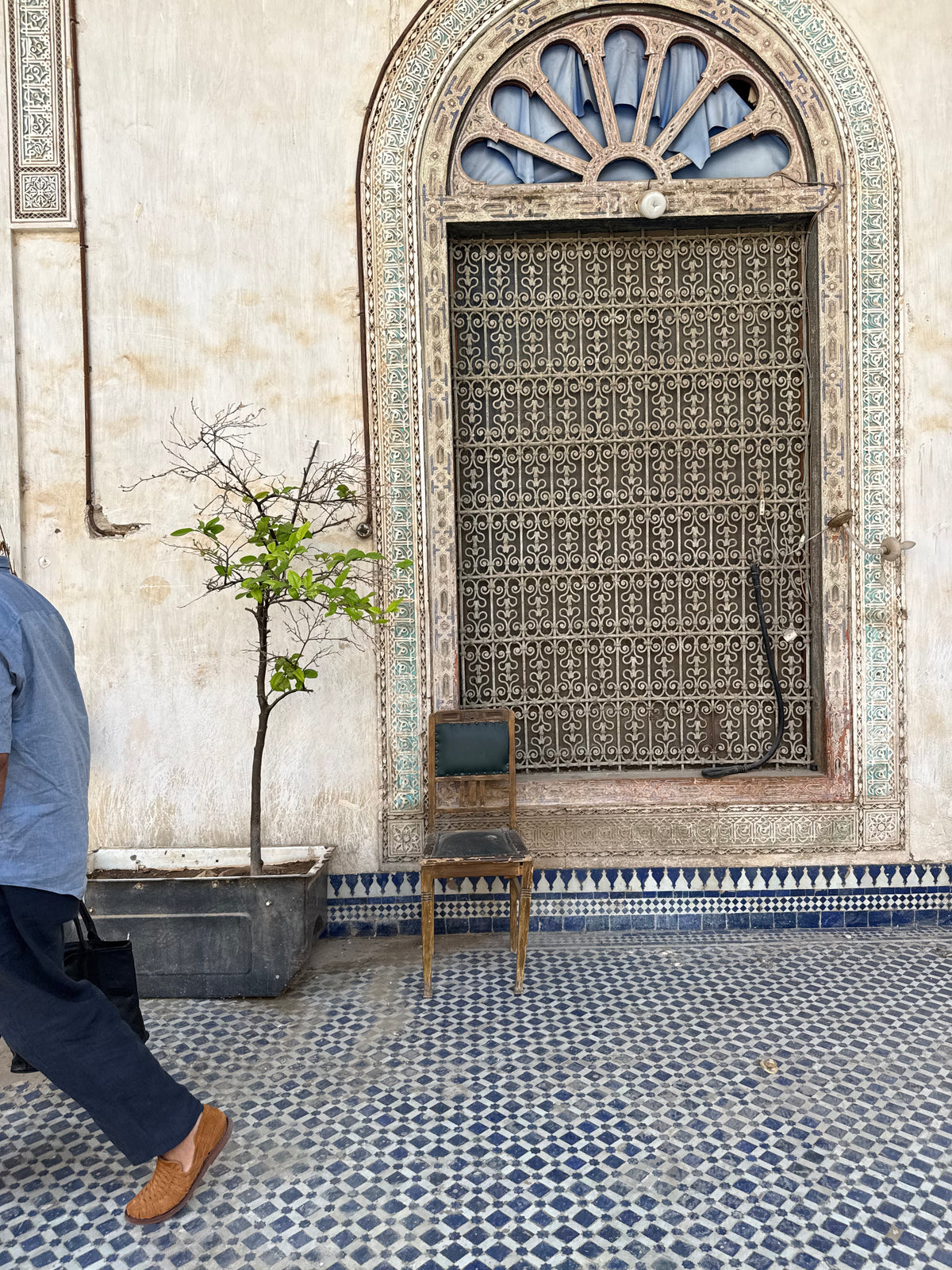 A person in a blue shirt and dark trousers walks past a traditional Moroccan-style window with a metal grille, a small wooden chair, and a potted tree on a blue-and-white patterned tile floor.
