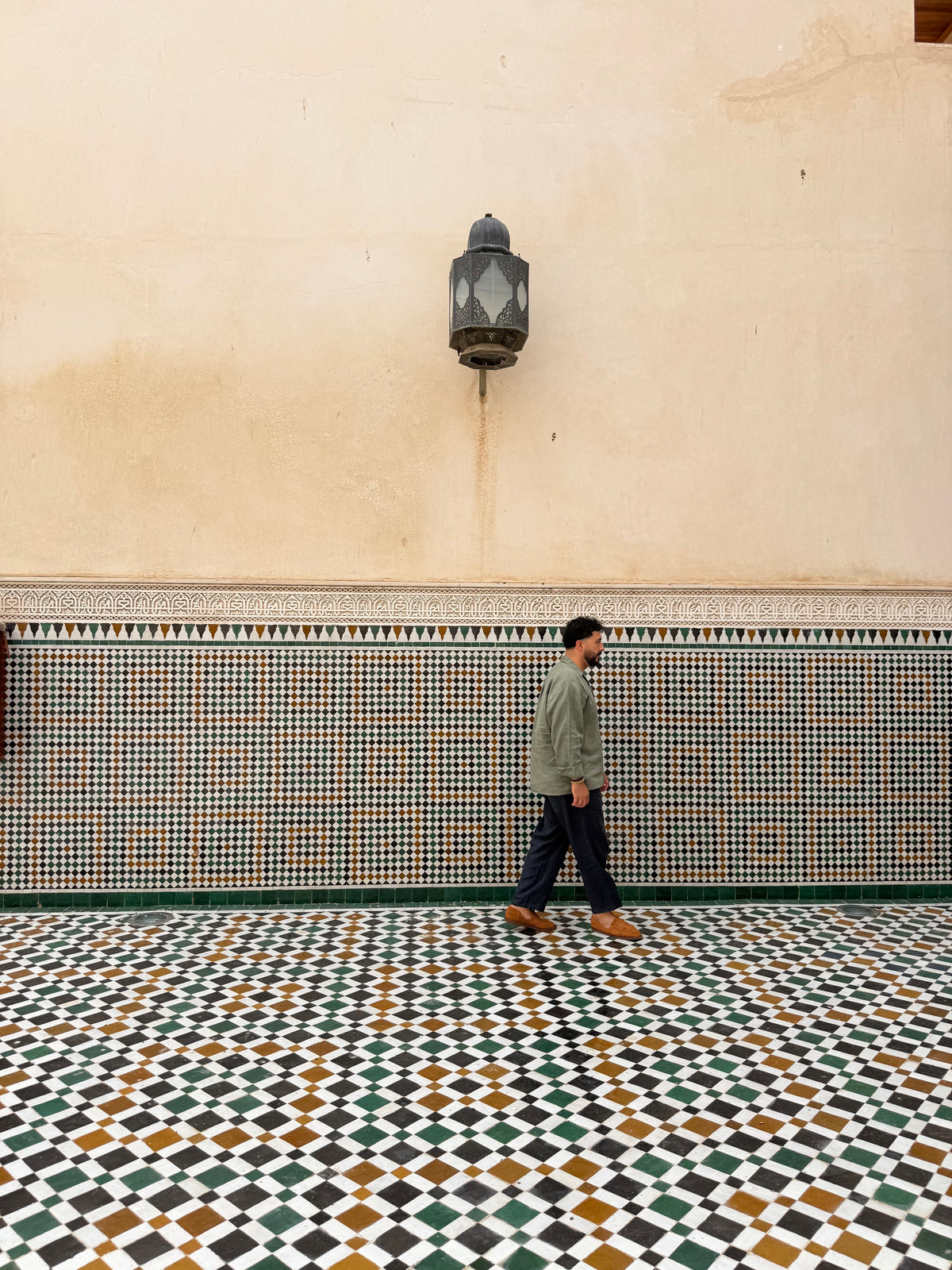 A man walks along a tiled corridor with a beige wall and a hanging lantern above him.