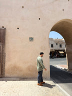 A man wearing a sage green long-sleeve shirt, dark gray pants, and brown boots stands in profile under a large beige stone archway.