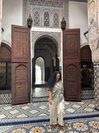 A woman with long curly black hair stands in a Moroccan-style courtyard with intricate tile floors, carved wooden doors, and geometric patterns. She is wearing a light sage green short-sleeve button-up shirt and off-white trousers.