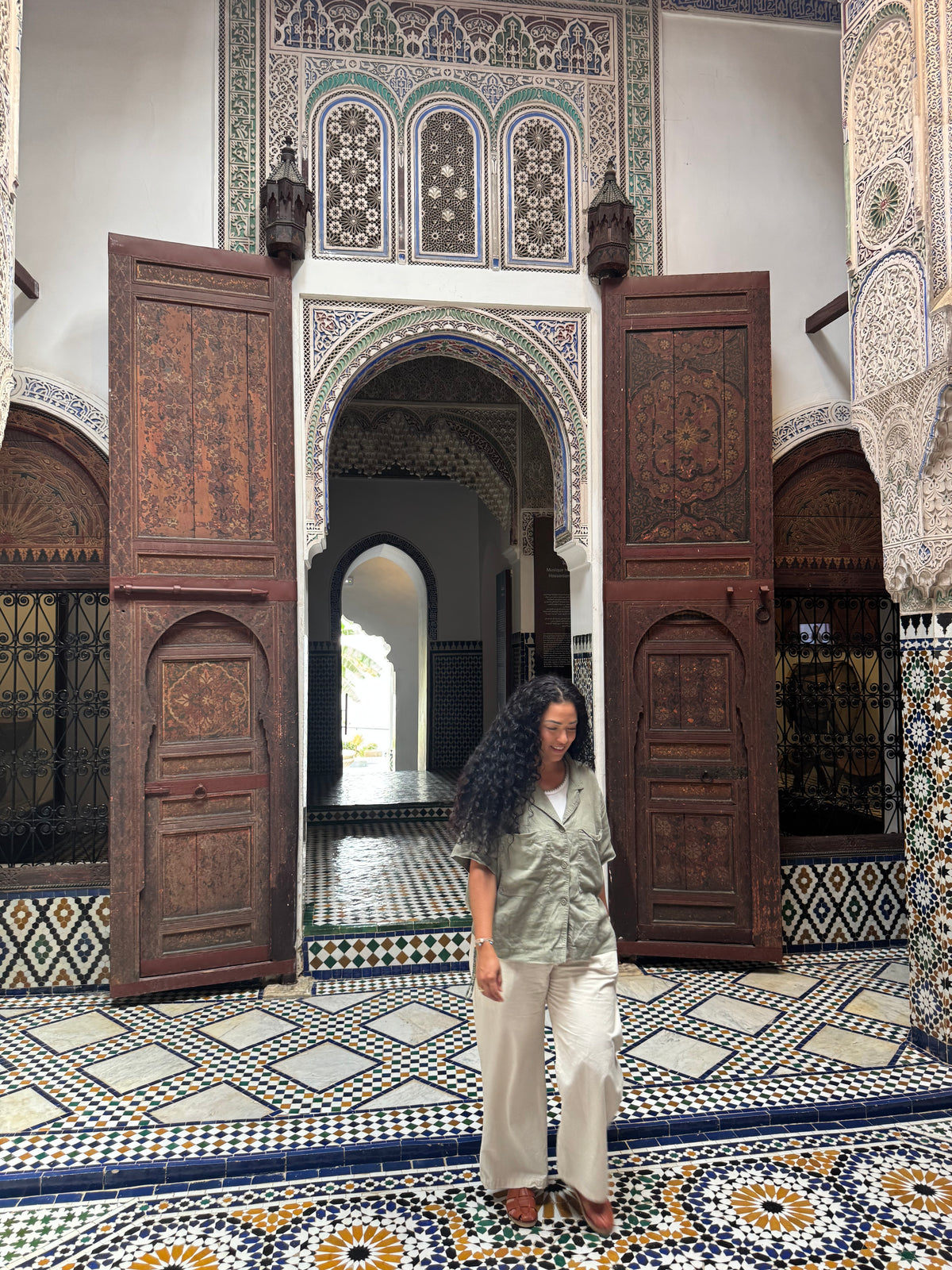 A woman with long curly black hair stands in a Moroccan-style courtyard with intricate tile floors, carved wooden doors, and geometric patterns. She is wearing a light sage green short-sleeve button-up shirt and off-white trousers.