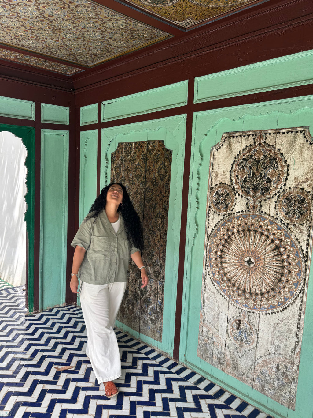 A woman wearing a sage green short-sleeve button-up shirt and white wide-leg pants walks inside a room with mint green walls, dark wood trim, patterned tapestries, and a blue-and-white chevron tile floor.