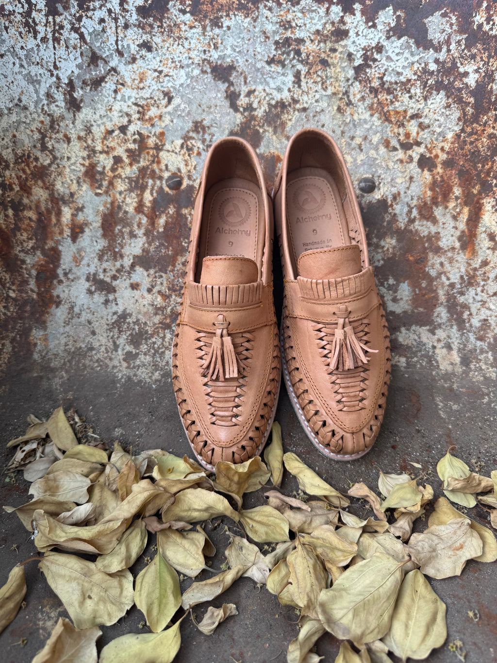Pair of brown loafers huaraches with tassels on a textured surface with dried leaves.