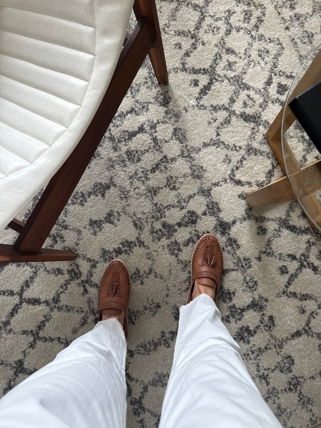 Person wearing brown loafer huaraches and white pants standing on a patterned carpet next to a chair.