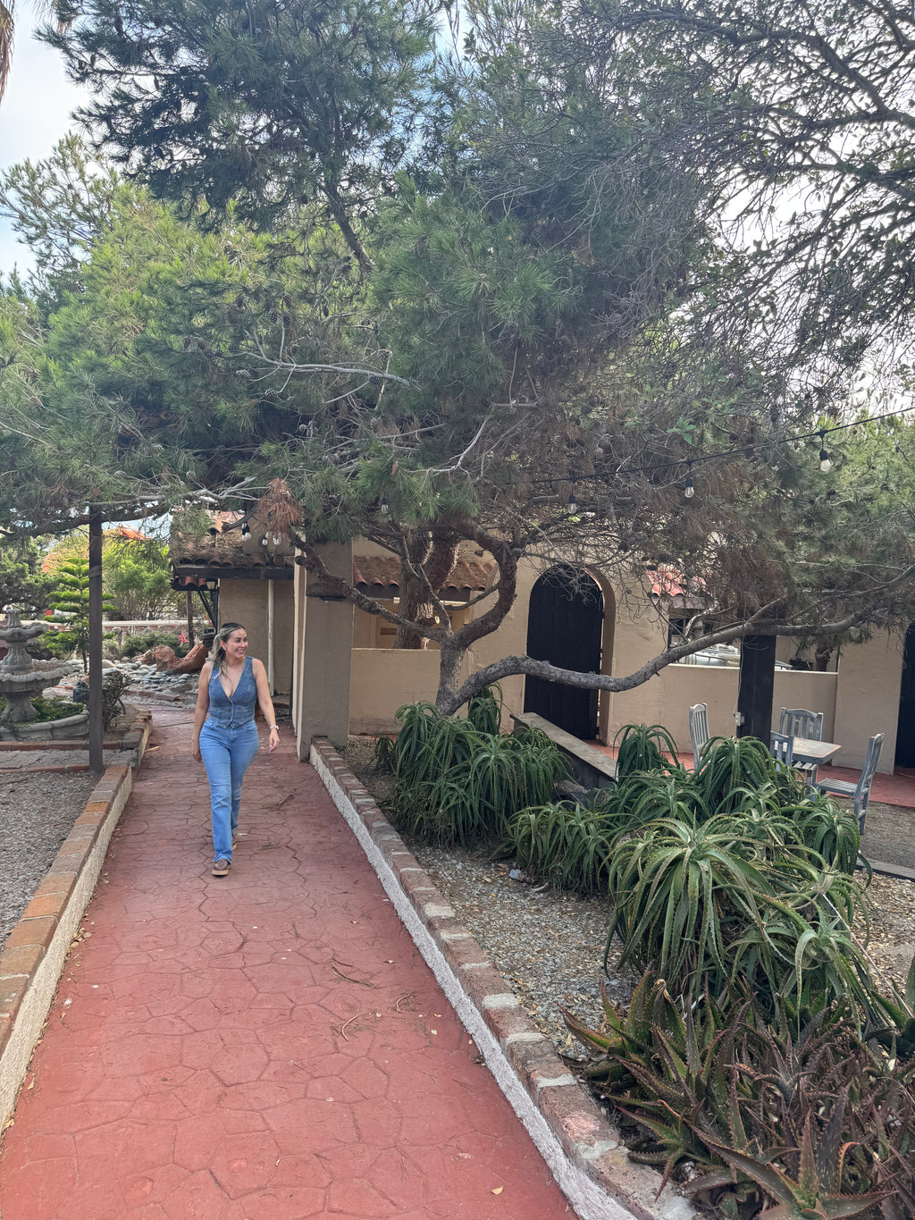 A woman wearing a blue denim overall jumpsuit walks along a red brick pathway in a landscaped garden courtyard with trees, plants, and a beige building in the background.
