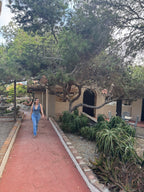 A woman wearing a blue denim overall jumpsuit walks along a red brick pathway in a landscaped garden courtyard with trees, plants, and a beige building in the background.