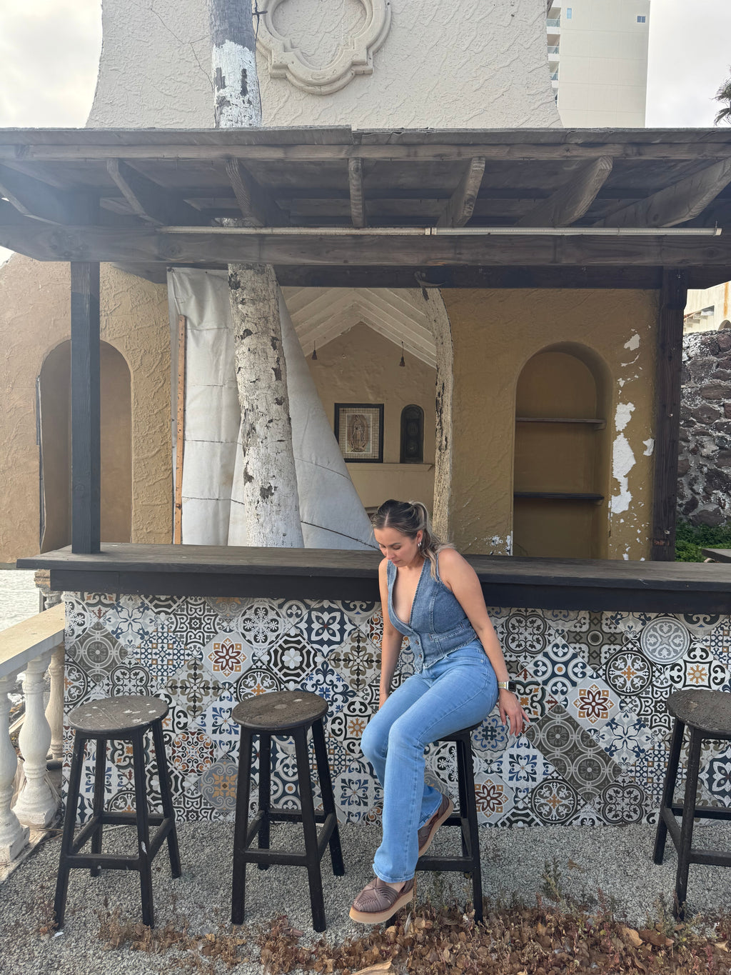 A woman wearing a blue denim jumpsuit sits on a black bar stool in front of a rustic tiled bar counter.