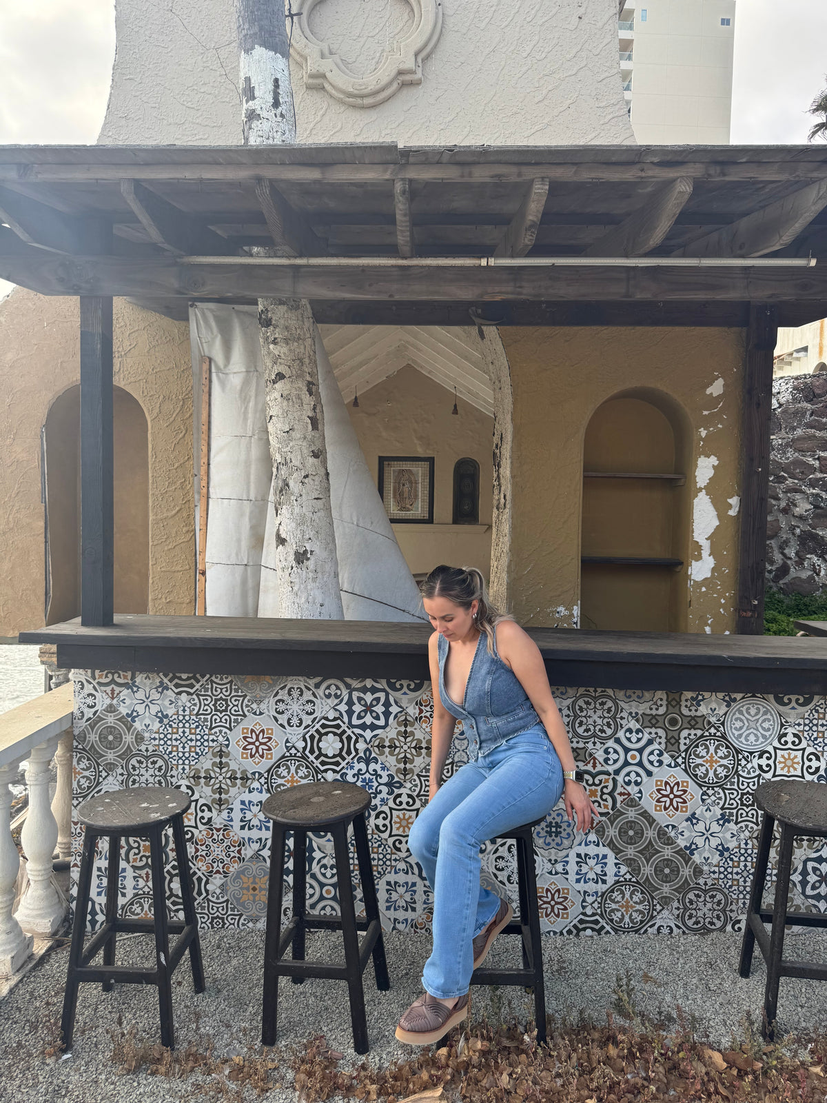 A woman wearing a blue denim jumpsuit sits on a black bar stool in front of a rustic tiled bar counter.