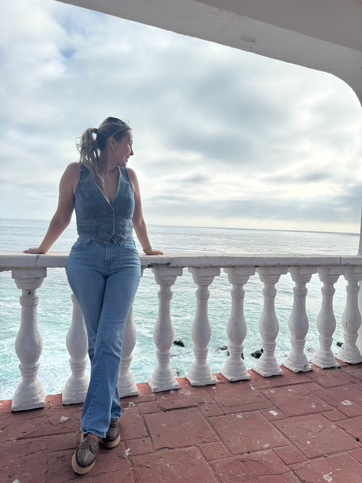 A woman stands on a balcony overlooking the ocean, wearing a sleeveless denim vest and light blue.jeans.