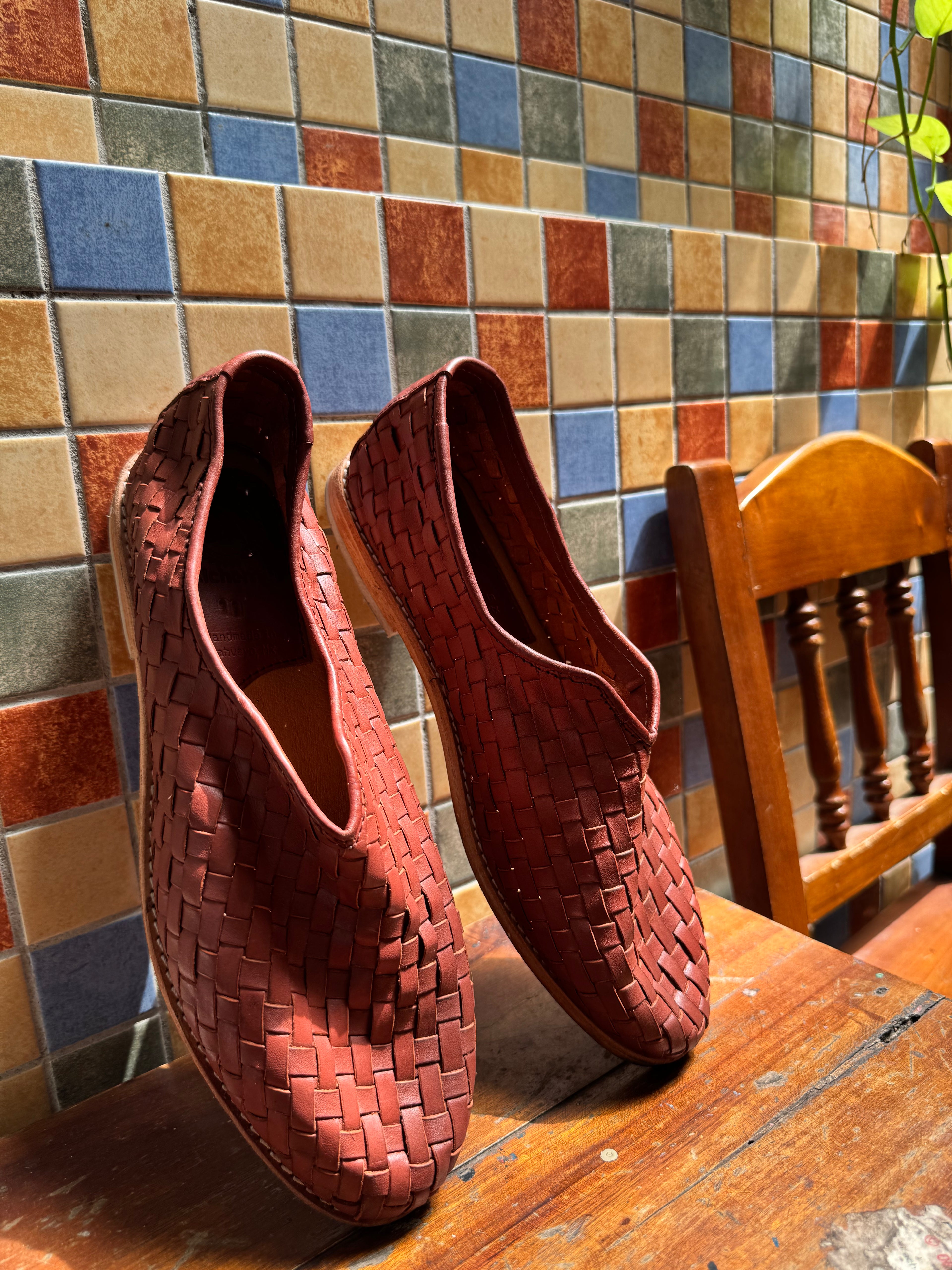Pair of woven red huaraches on a wooden surface with a multicolored tiled wall in the background