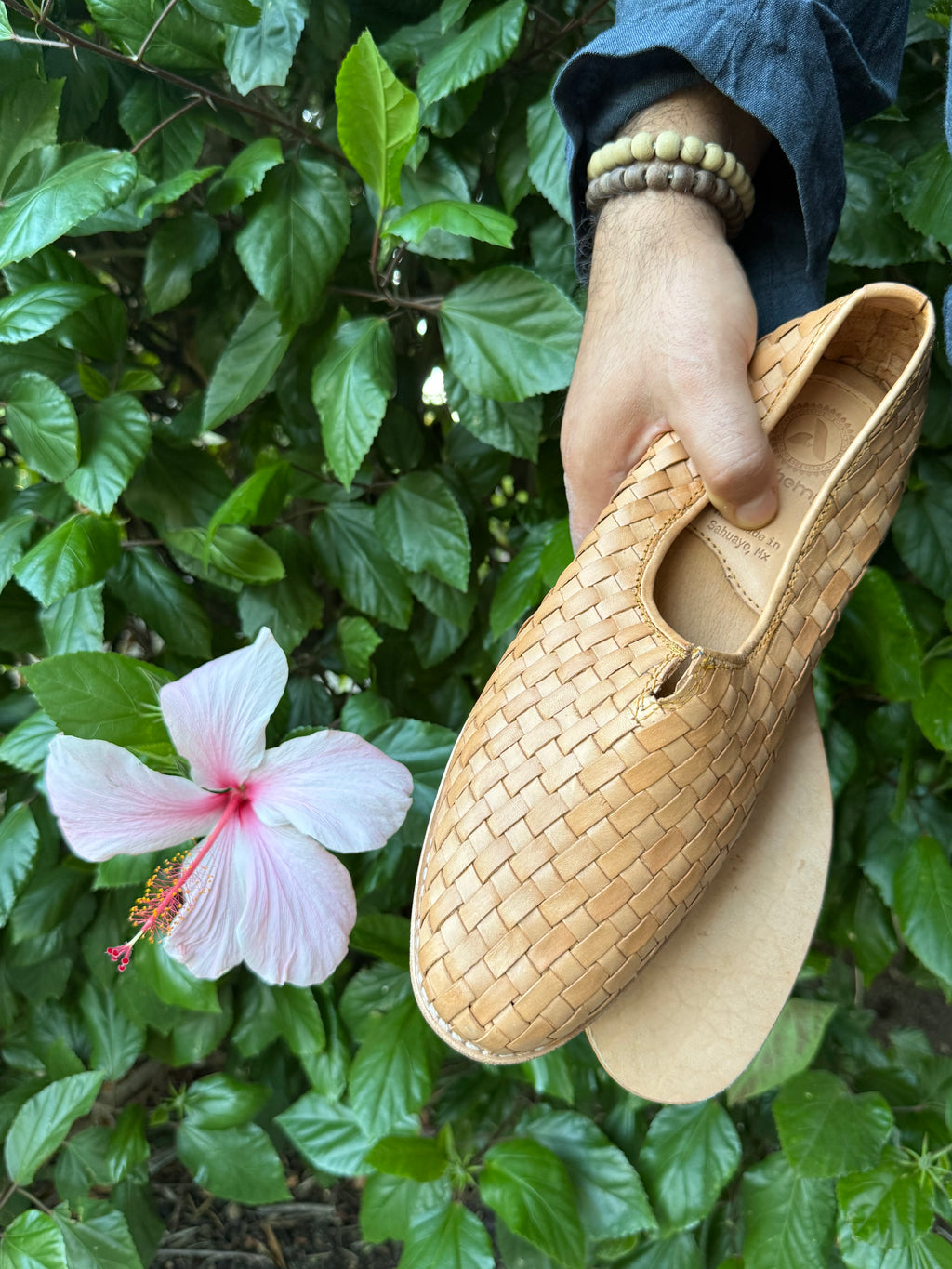 Hand holding a woven huaraches with a pink flower and green leaves in the background.