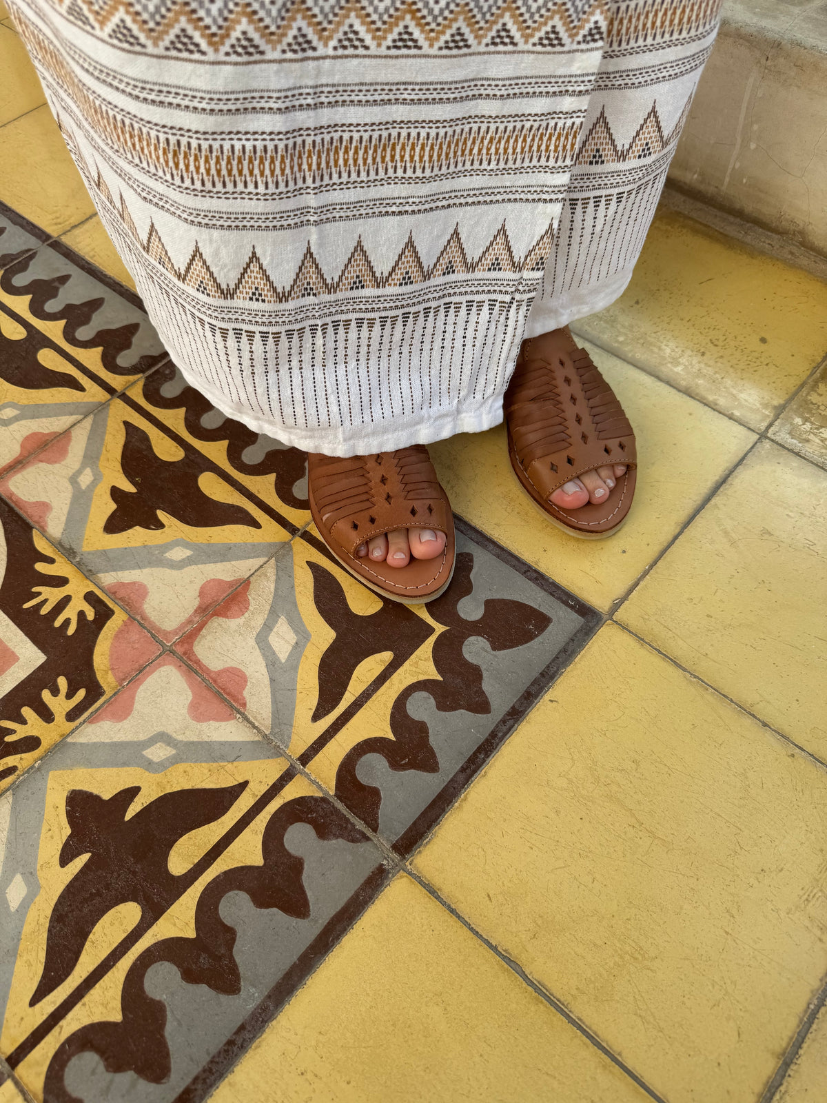 A person wearing a white knit skirt with brown geometric patterns and huaraches, standing on a yellow tiled floor with a decorative brown, gray, and yellow patterned rug.